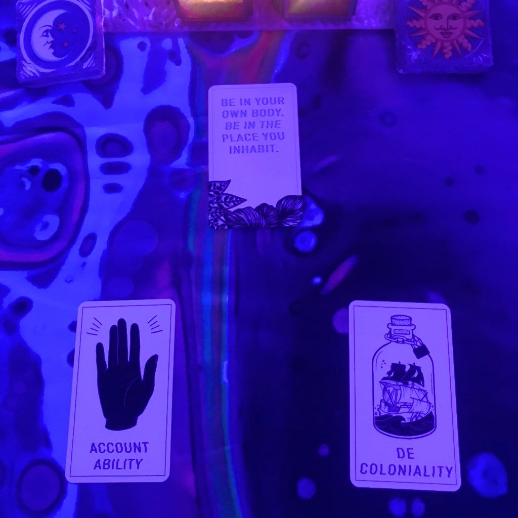 Three cards under blacklight, At the top, a card with the words "Be in your own body. Be in the place you inhabit." On the bottom left, a hand and the word Accountability. On the bottom right, a ship in a bottle with a lock on it, and the word Decoloniality.