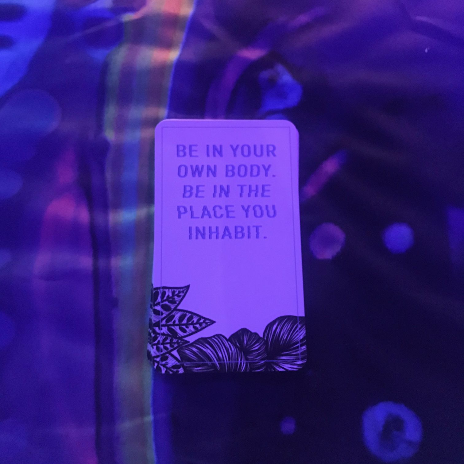 A card on a colorful reading cloth, under black light, with the words "Be in your own body. Be in the place you inhabit."