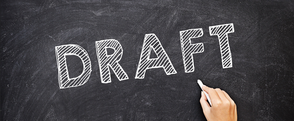 The word Draft, written in white chalk on a blackboard.  On the right, a hand holding a piece of white chalk.